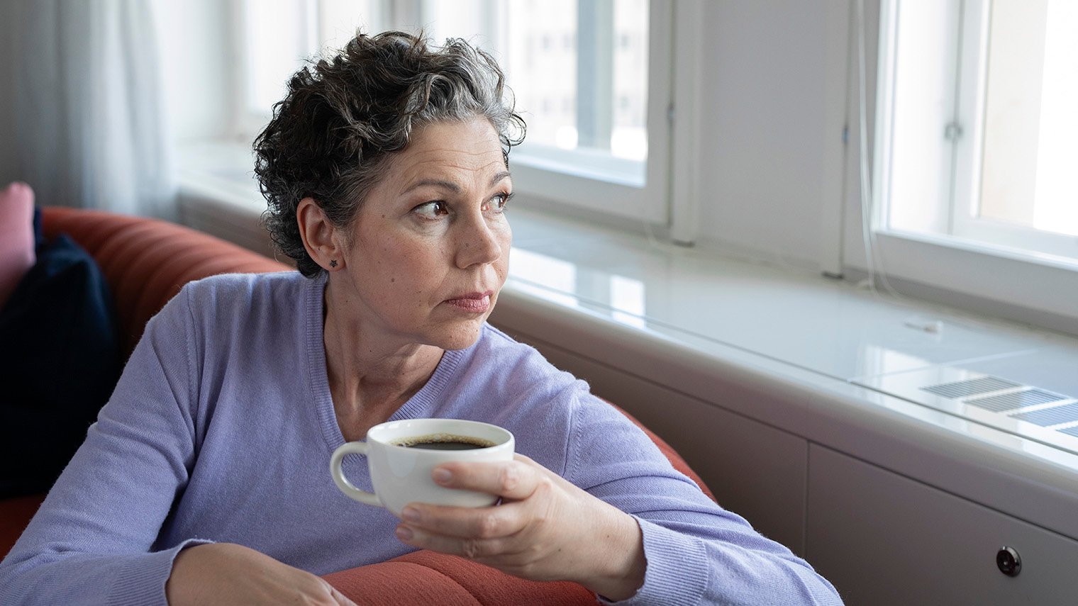 Vakava nainen katsoo ikkunasta ulos kahvikuppi kädessään.